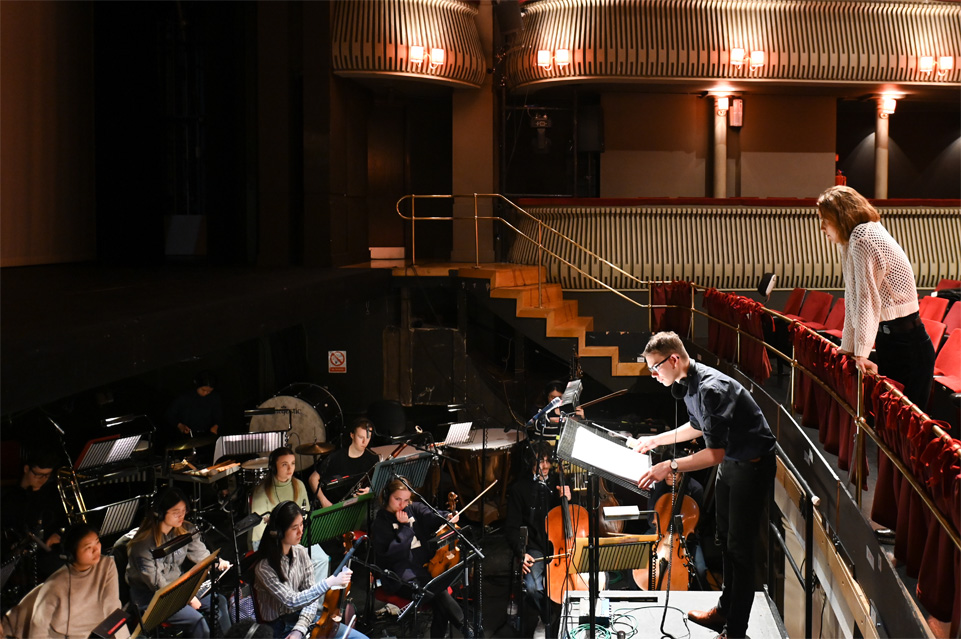 A female student looking over an orchestra pit, where a male conductor and a group of students holding music instruments rehearsing the music she composed.